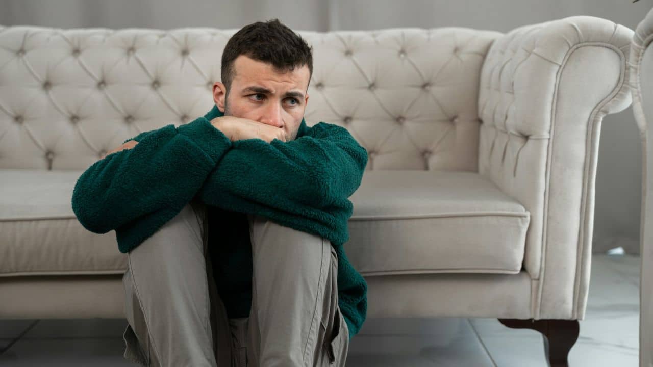 A man in a green sweater and light pants sits on the floor, looking worried.