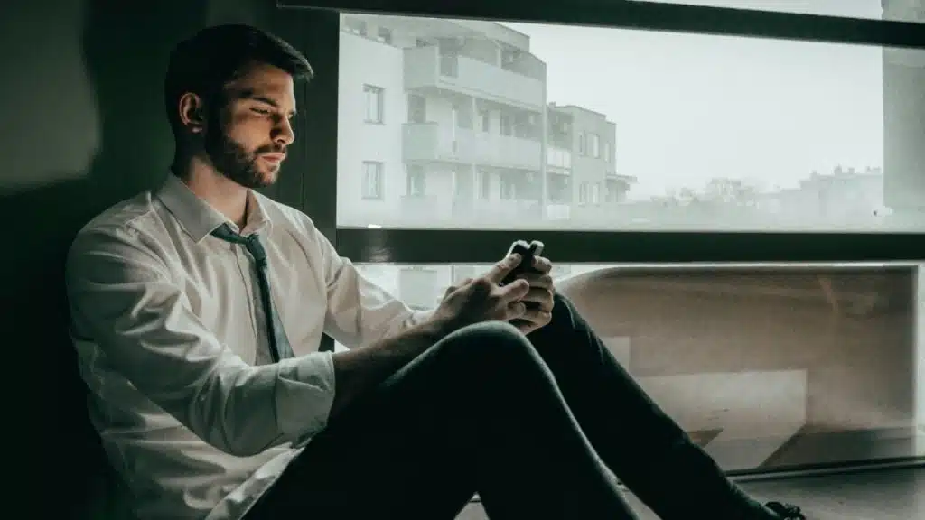 A pensive man sits by a window, looking at his phone.