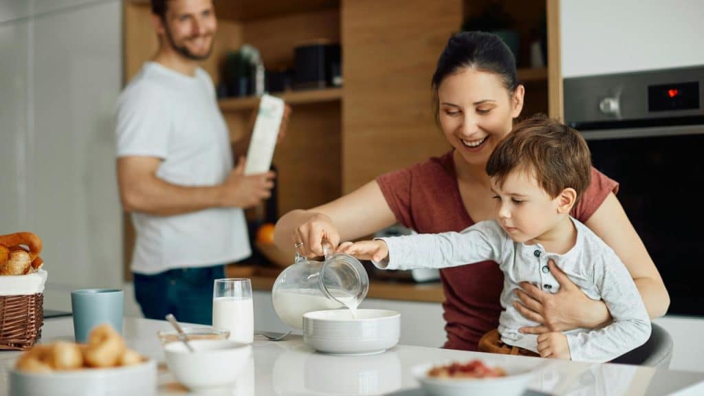 A mother pouring milk into her toddler’s cereal bowl while the father holds a cereal box.