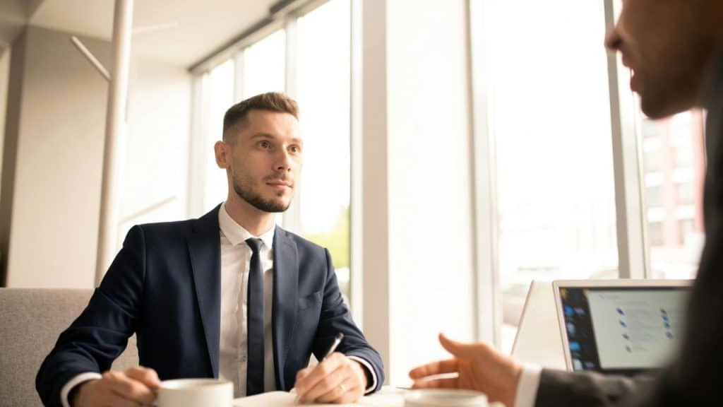 A man in a navy suit and tie sits at a table, looking forward, while holding a pen and a cup.