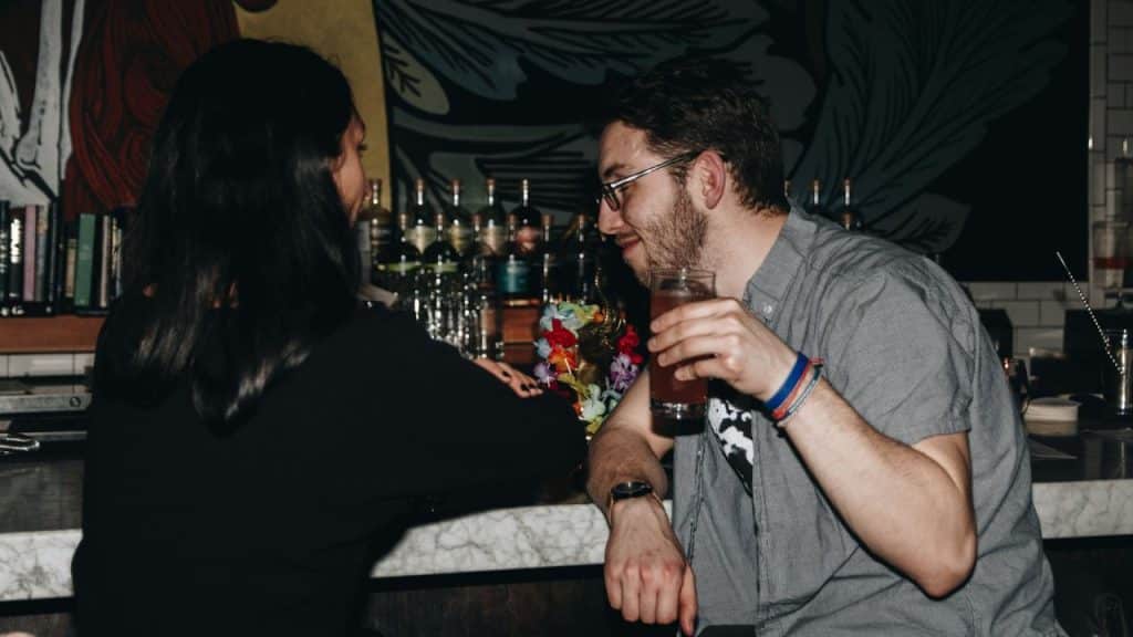 A man wearing glasses holds a drink and talks to a woman at a bar.