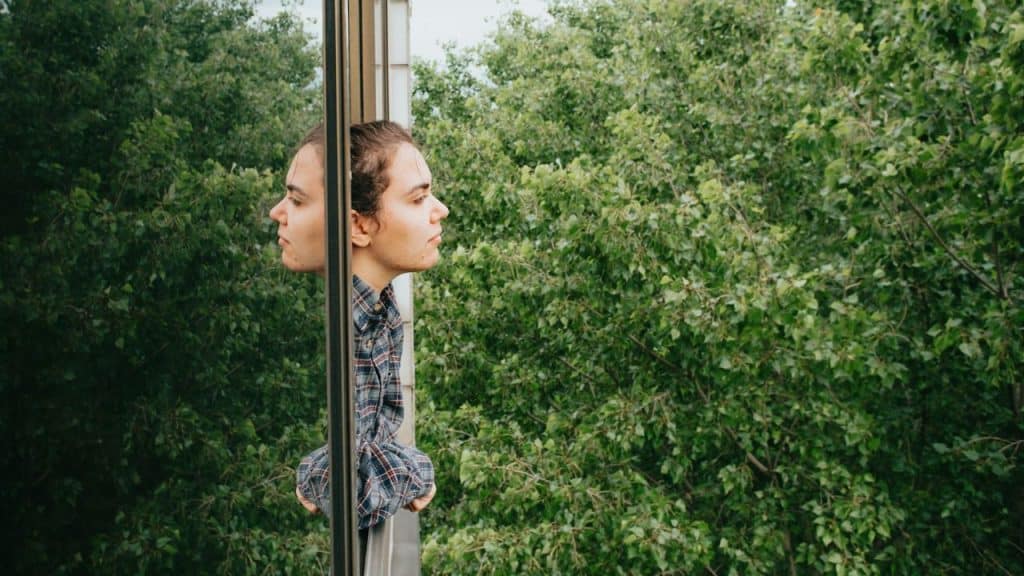 A woman looks out a window, with his reflection visible on the glass.