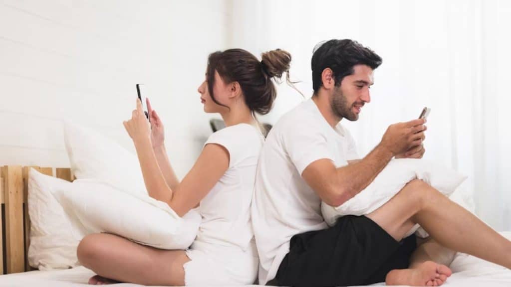 Couple sitting back to back on a bed, both focused on their phones, suggesting emotional distance