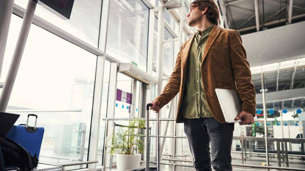 A man in an airport terminal holding a laptop and pulling a rolling suitcase.