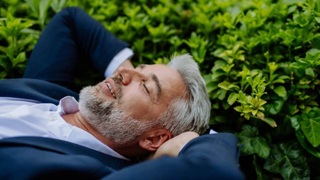 A man in a suit resting on green bushes.