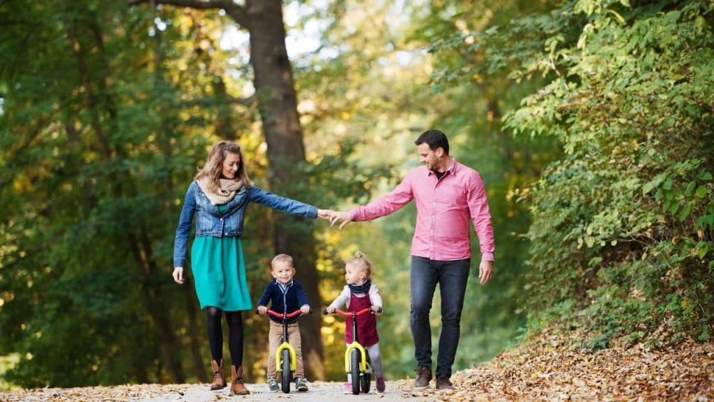 A mother and father holding hands over two children riding balance bikes.
