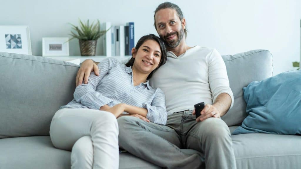 A happy couple is seated on a couch, with the man holding a remote control.