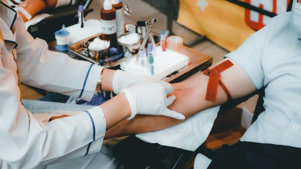 A medical professional in white gloves draws blood from a person's arm, with vials and medical supplies in the background.