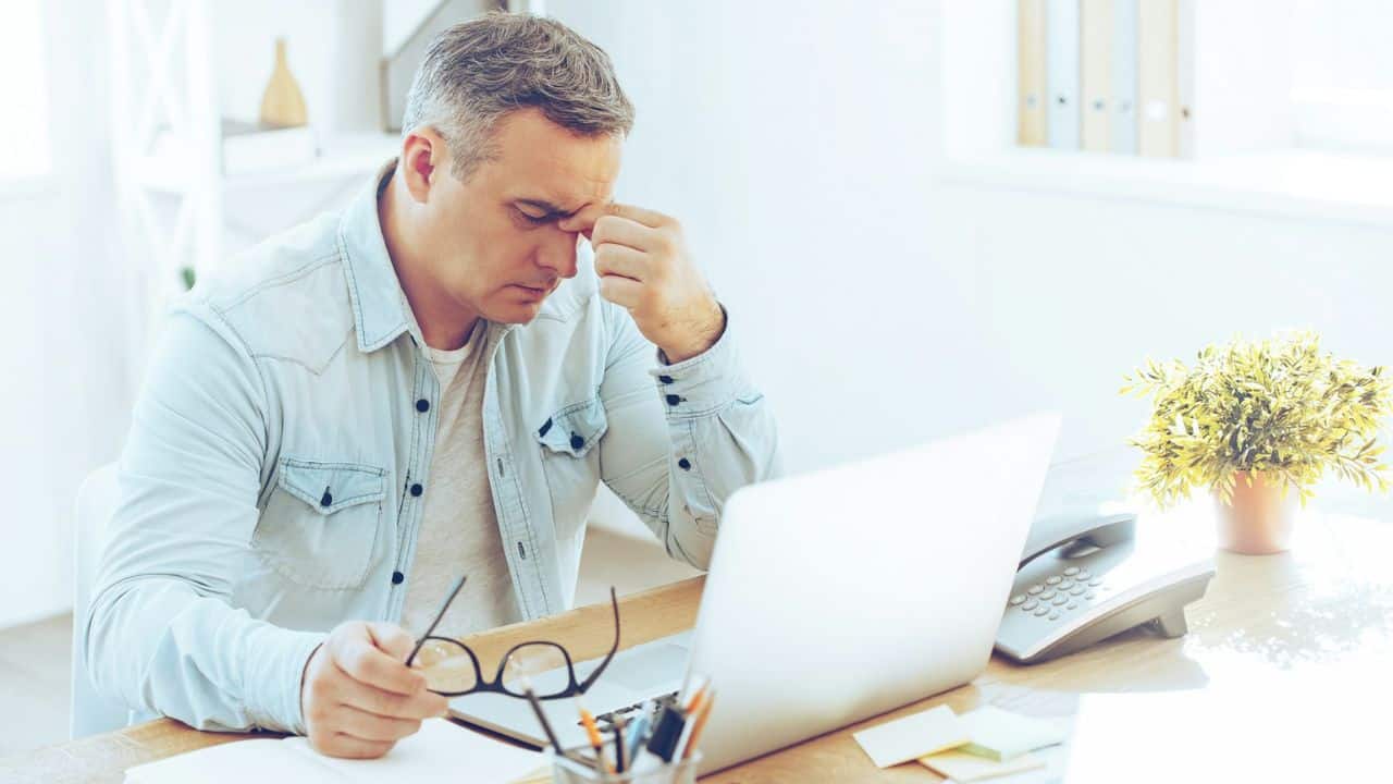 A man in a denim shirt sits at a desk, rubbing his forehead.