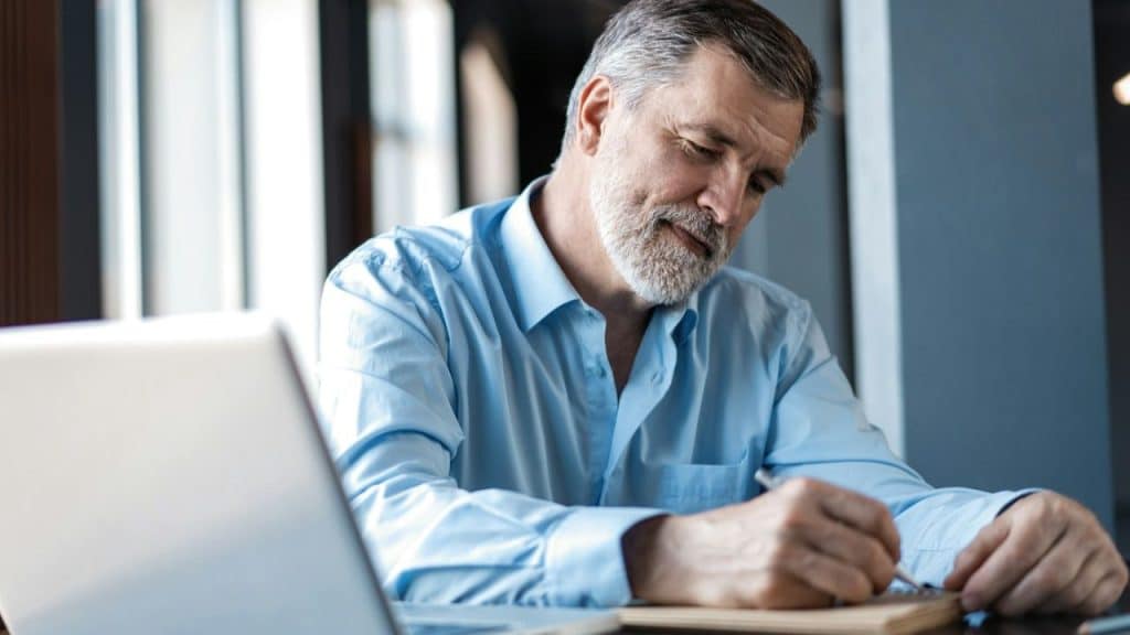 A smiling, gray-haired man in a light blue shirt writes in a notebook.