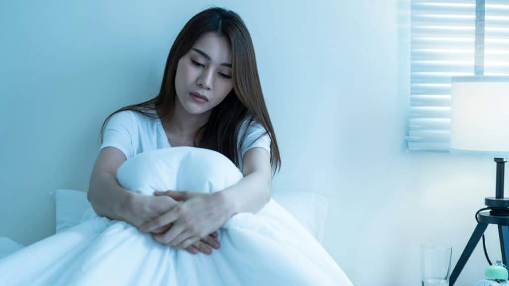 A somber woman sits on a bed, hugging a white pillow.