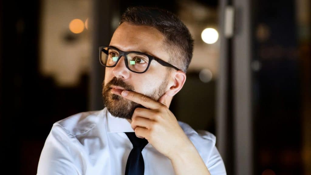 A bearded man wearing glasses and a tie thoughtfully looks to the side.