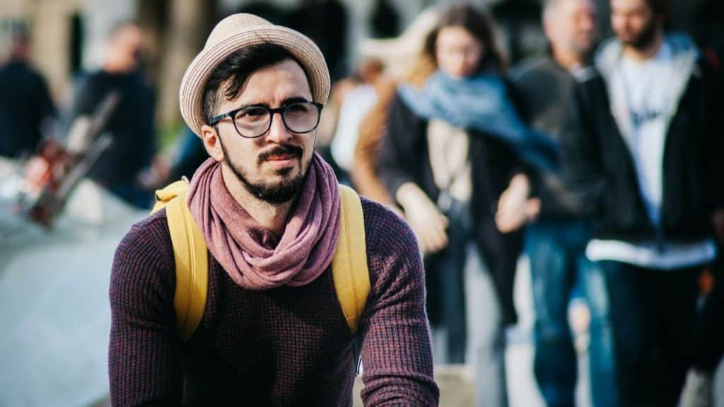 A person wearing glasses, a hat, and a scarf looks off to the side, with blurry figures in the background.