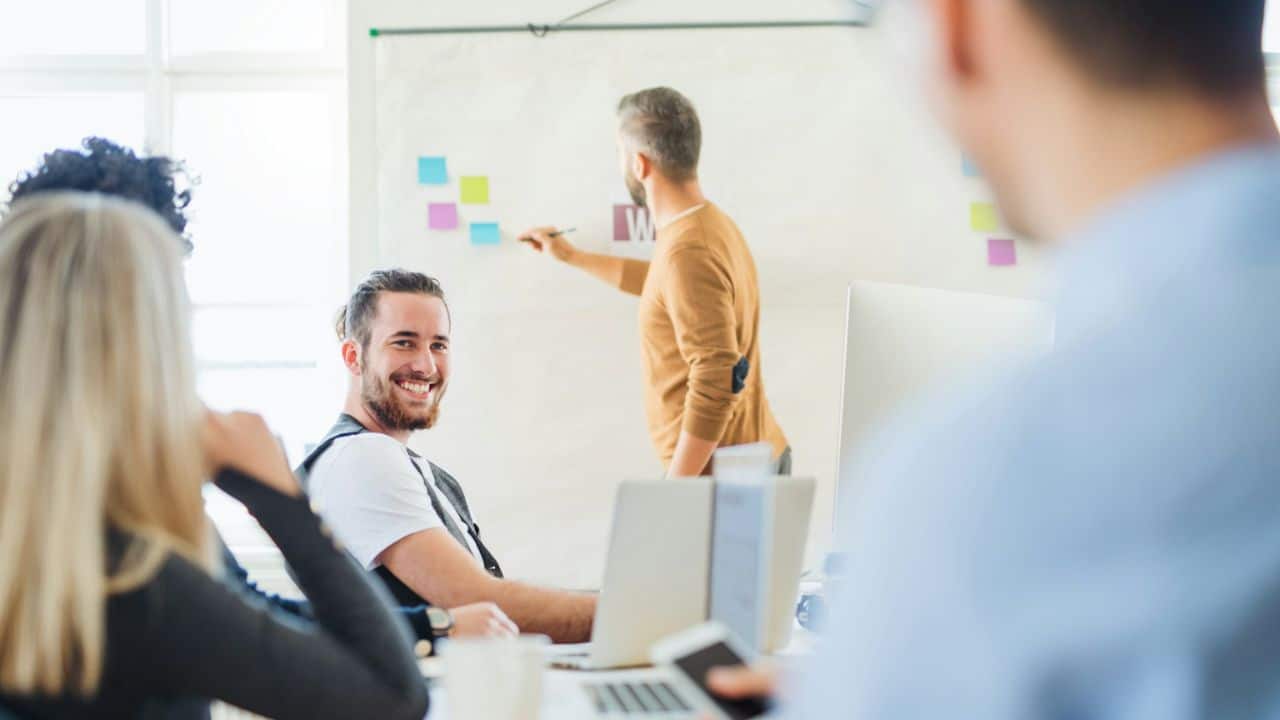 A man with a beard smiles at the camera while another man writes on a whiteboard behind him.