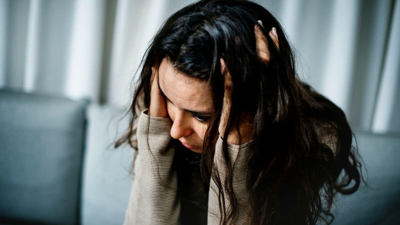 A woman with dark, long hair holds her head in her hands, looking down with a distressed expression.