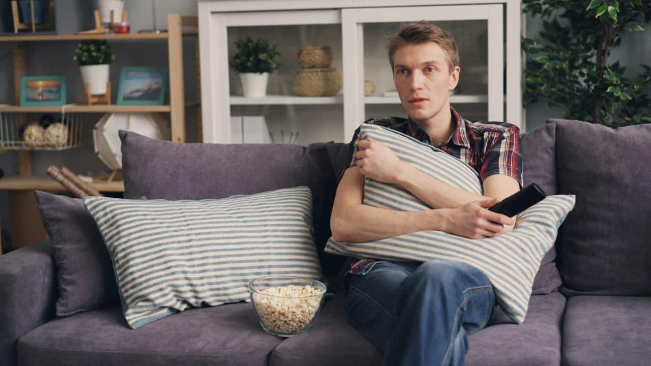 A man on a sofa holds a pillow, watching TV with a bowl of popcorn nearby.