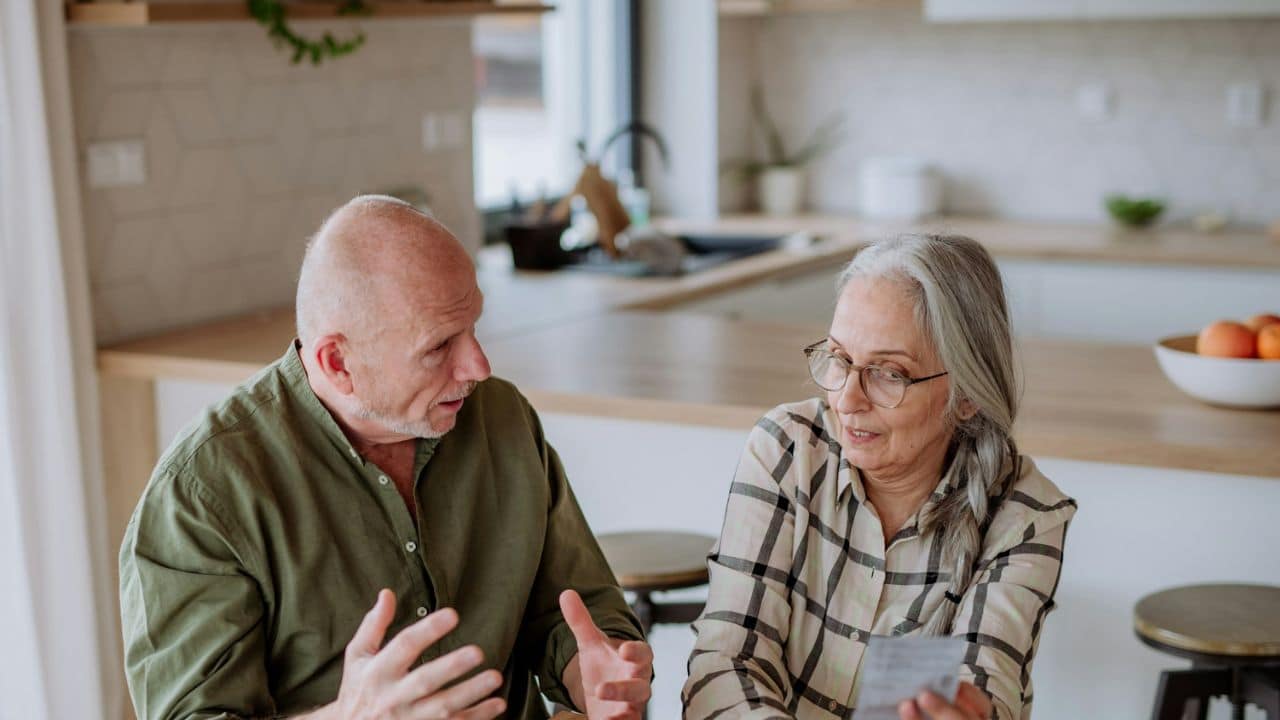 A mature couple sits at a kitchen table, engaged in a serious conversation..