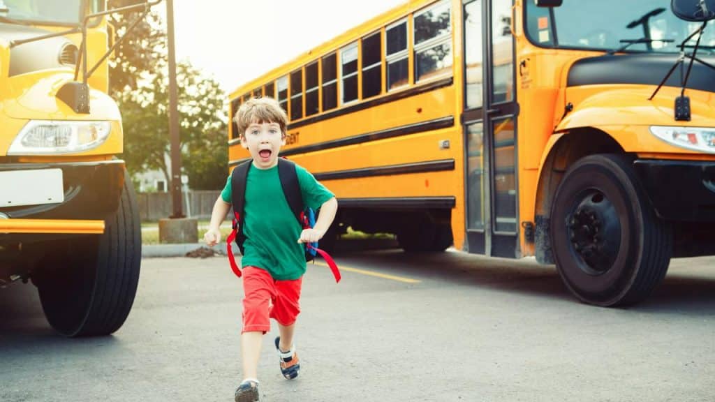 A boy running past a yellow school bus with a backpack.