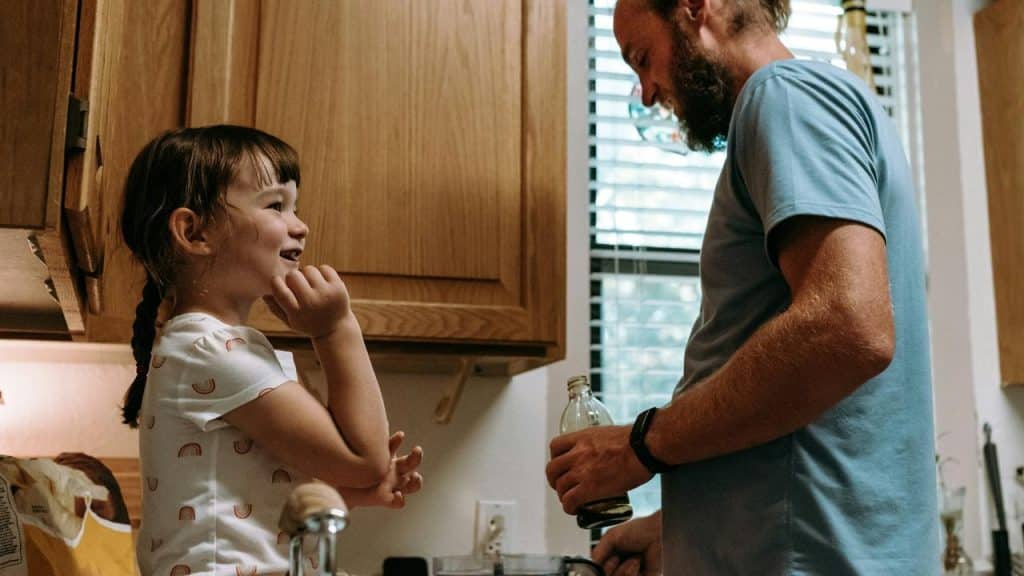 A girl smiling at a man pouring oil in a kitchen.