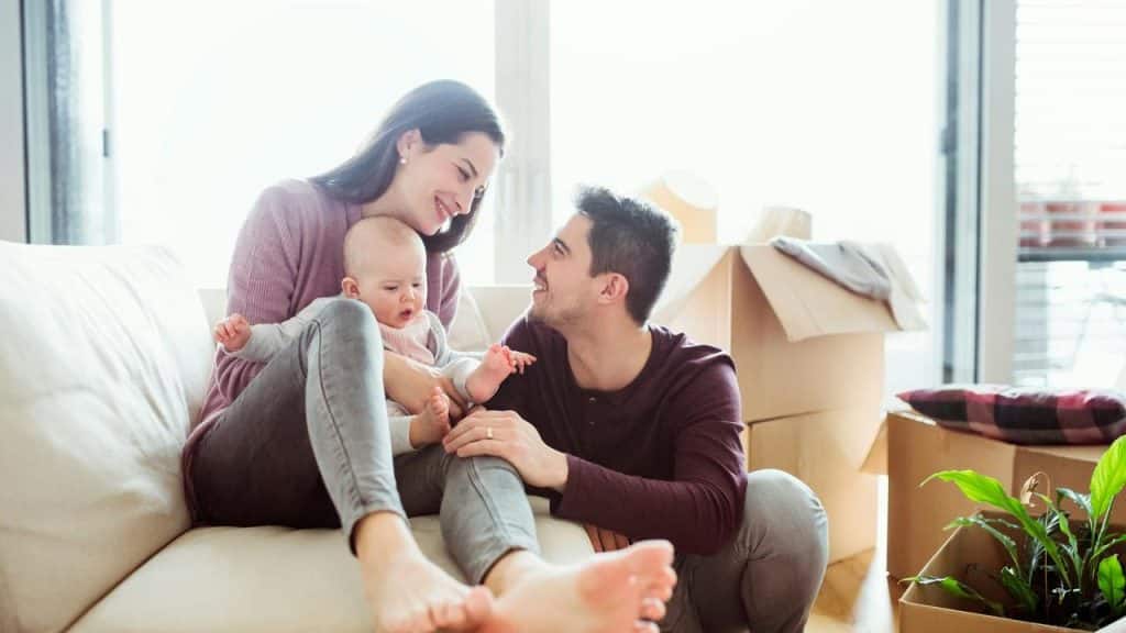 A smiling couple with their baby on a couch surrounded by moving boxes in a sunlit living room.