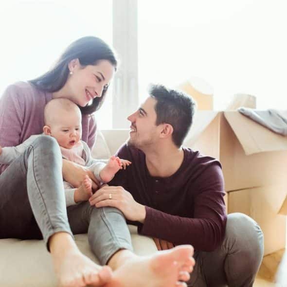 A smiling couple with their baby on a couch surrounded by moving boxes in a sunlit living room.