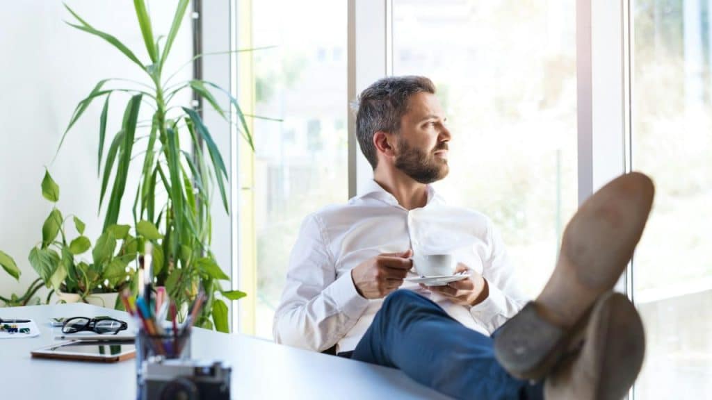 A relaxed man in a white shirt and jeans with his feet on a desk, sips coffee.