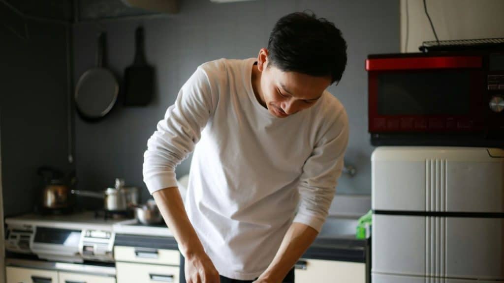 A man in a white long-sleeve shirt bends over in a kitchen, looking down.