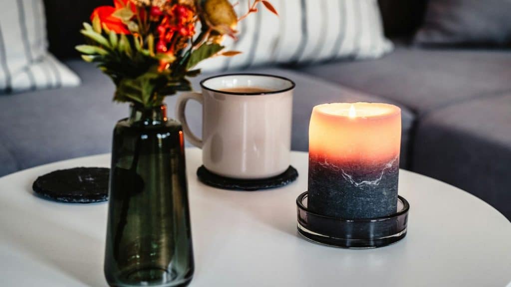 A lit, two-toned scented candle sits on a white table with a mug and vase.