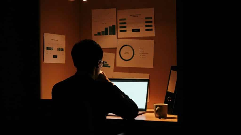 A man works at a laptop in a dimly lit room, with charts and graphs on the wall.