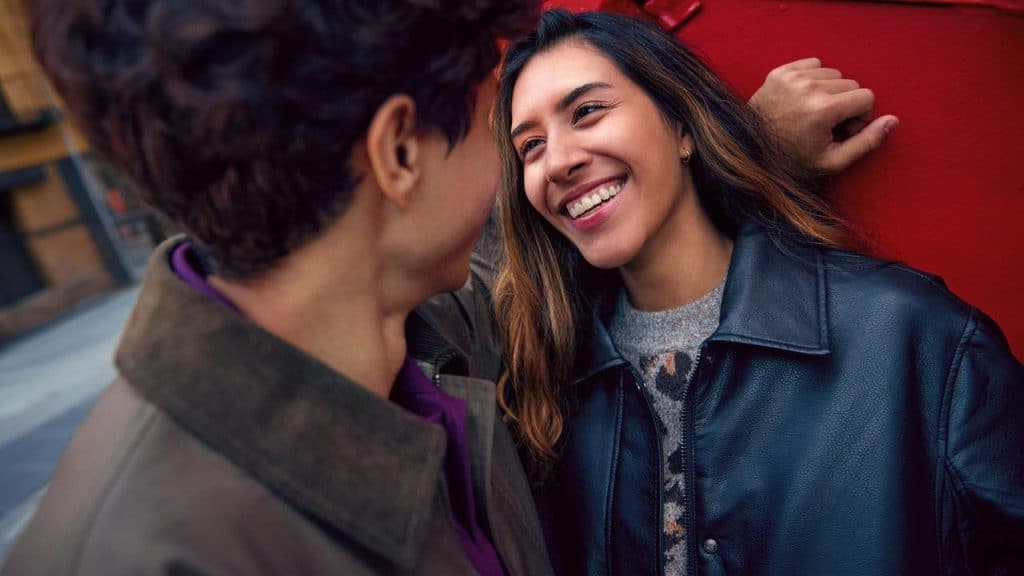 A smiling woman leaning against a red wall facing her companion.