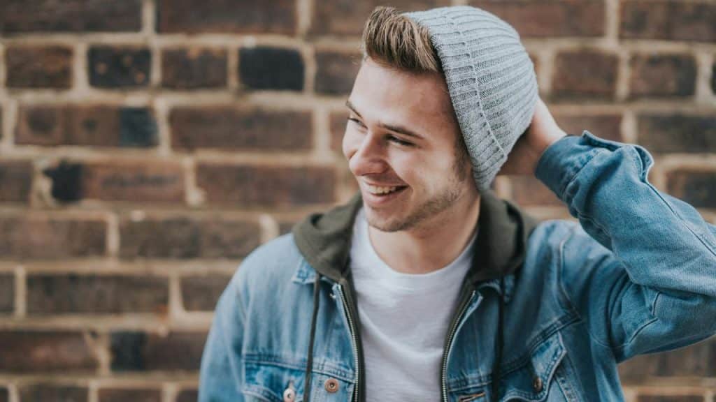 A smiling man wearing a beanie and denim jacket against a brick wall.