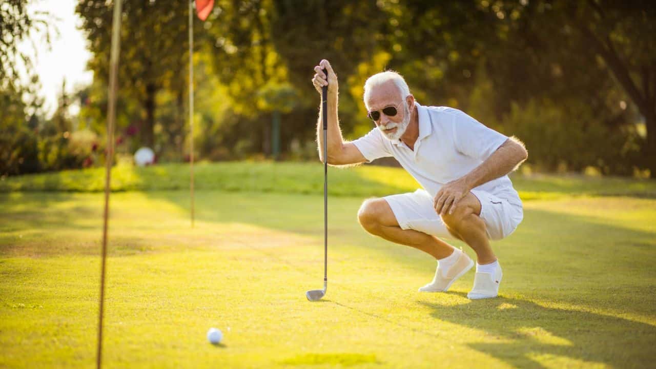 A white-bearded man in sunglasses crouches on a golf course, holding a club.