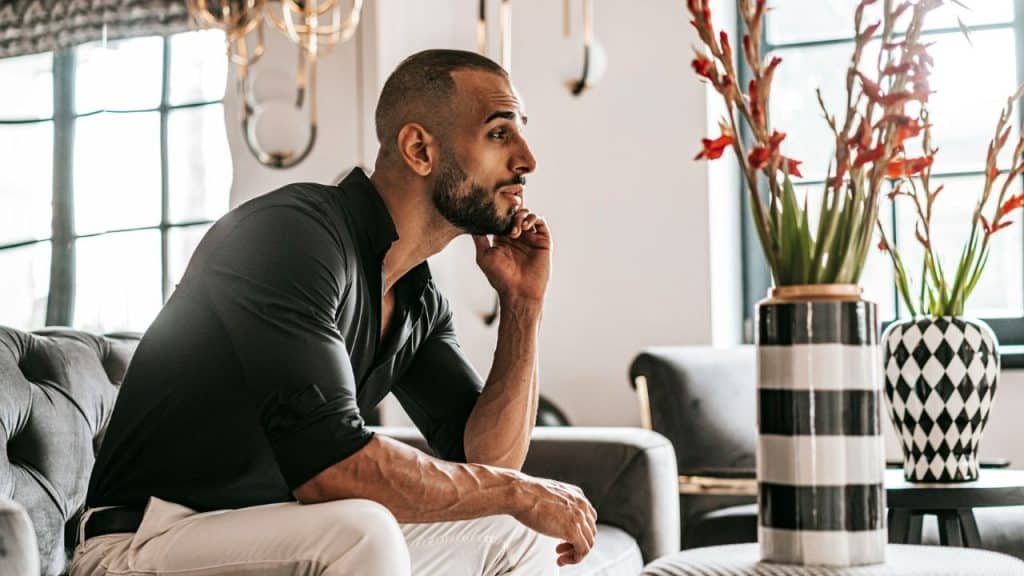 A thoughtful man with a beard sits on a sofa next to a vase with red flowers.