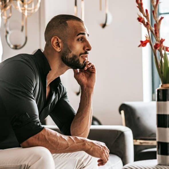 A thoughtful man with a beard sits on a sofa next to a vase with red flowers.