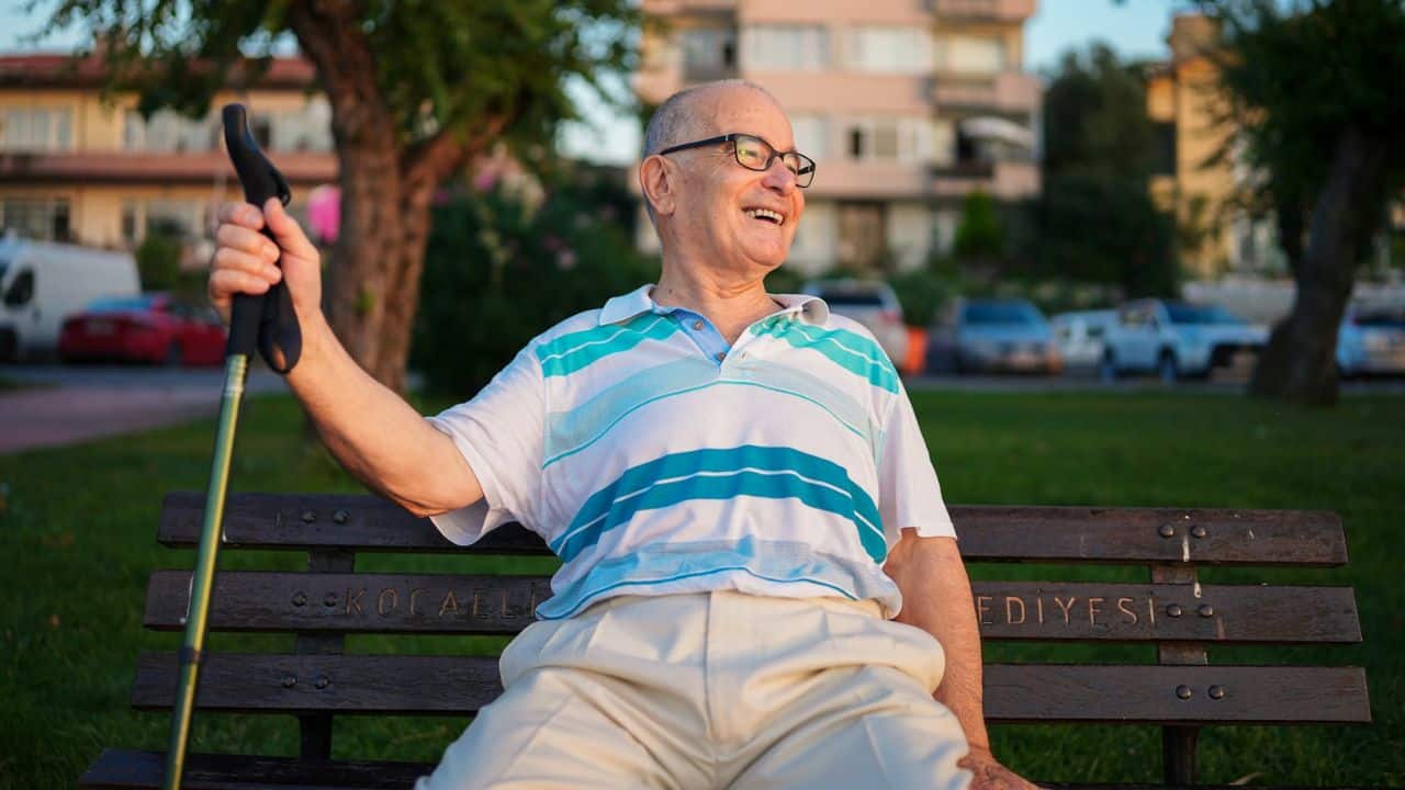 A smiling elderly man with glasses and a striped polo sits on a bench, holding a cane.