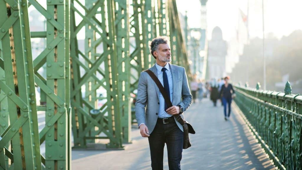 A man in a suit jacket and jeans walks across a green metal bridge.