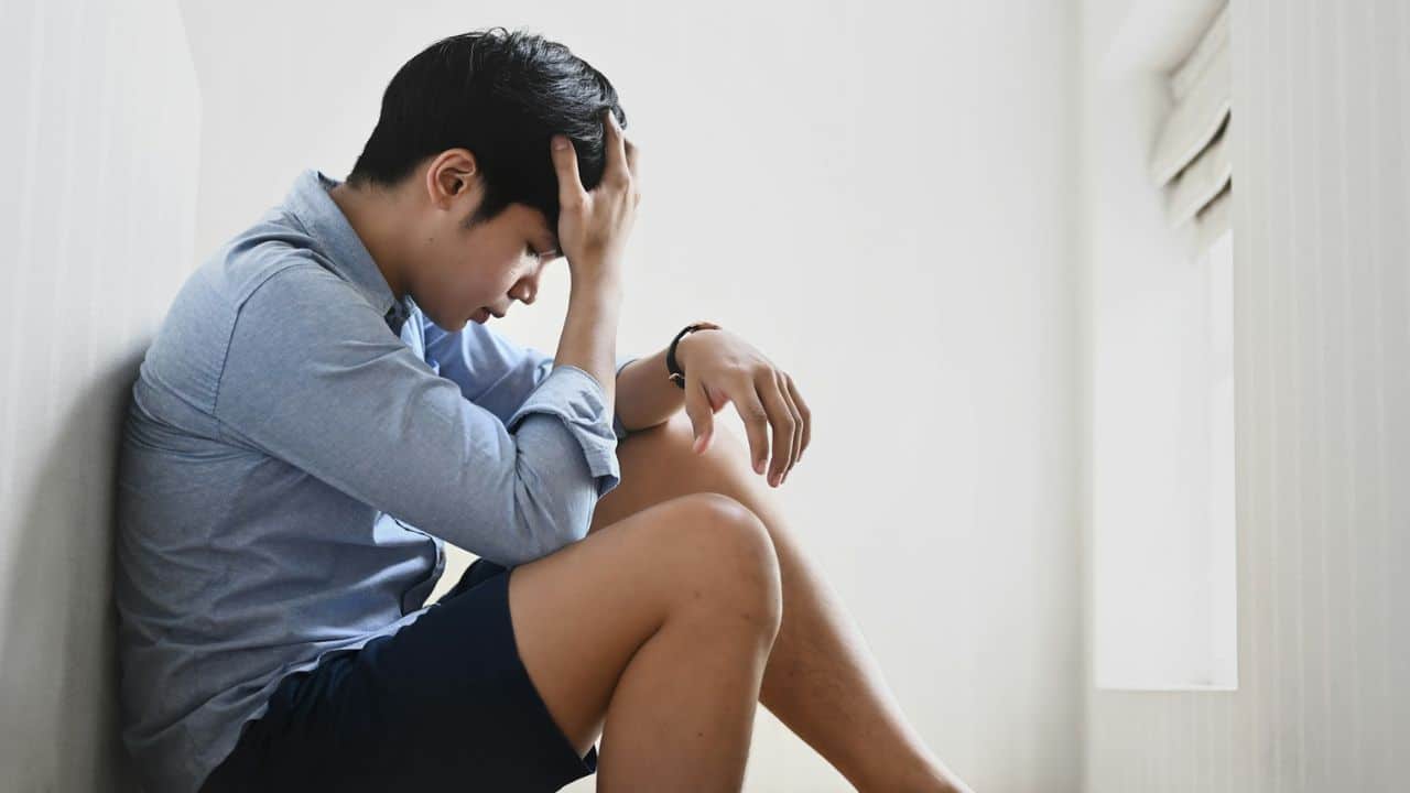 A young man sits on the floor with his head in his hands, looking distressed.