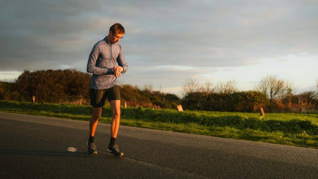 A man in athletic wear checks his watch while walking on a paved road.