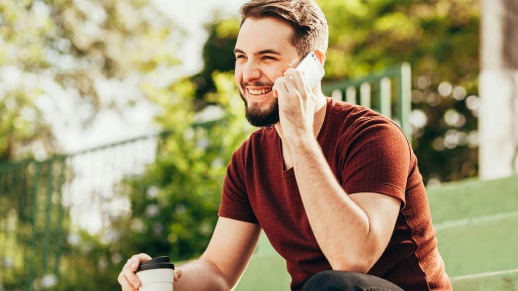 A bearded man smiling while talking on his phone and holding a coffee cup.