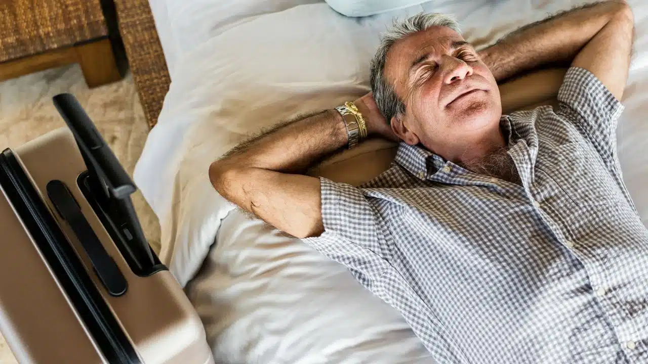A contented older man relaxes on a bed with hands behind his head.