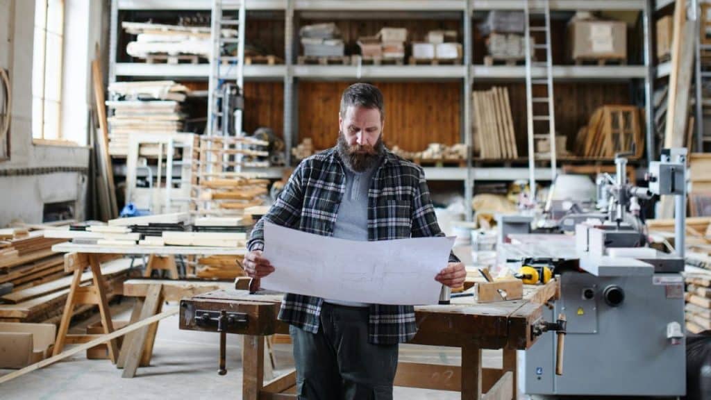 A bearded man in a plaid shirt examines blueprints in a woodworking shop.