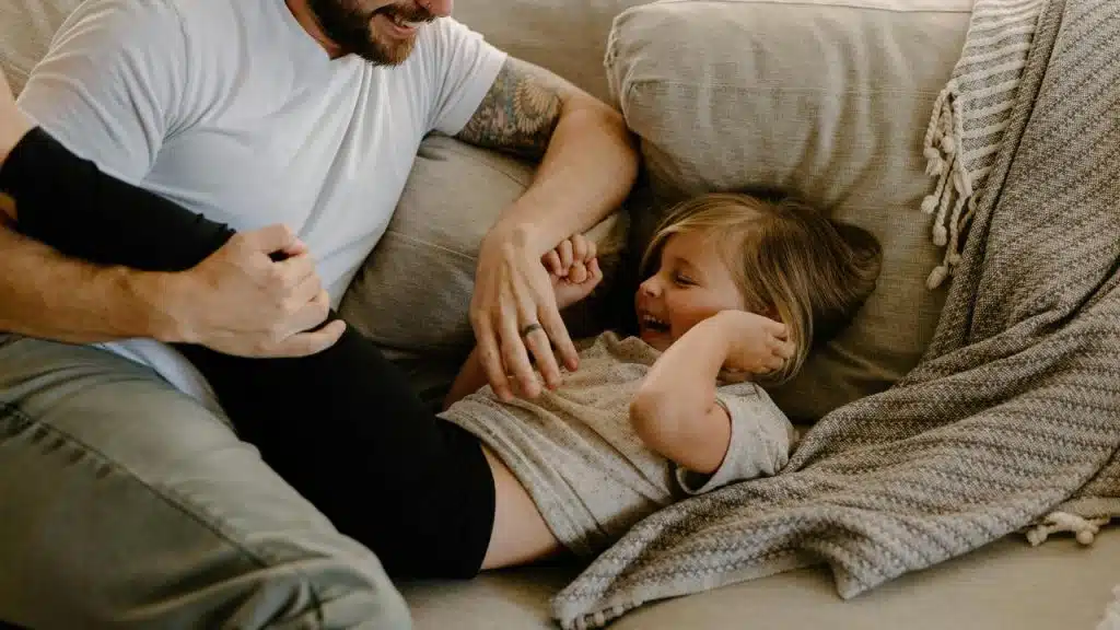 A man tickling his laughing young daughter as she lies on a couch.