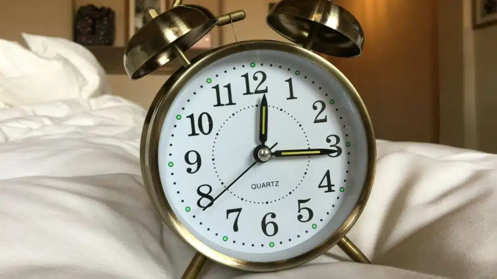 A classic gold alarm clock sits on a white bedspread, set to 3:00.