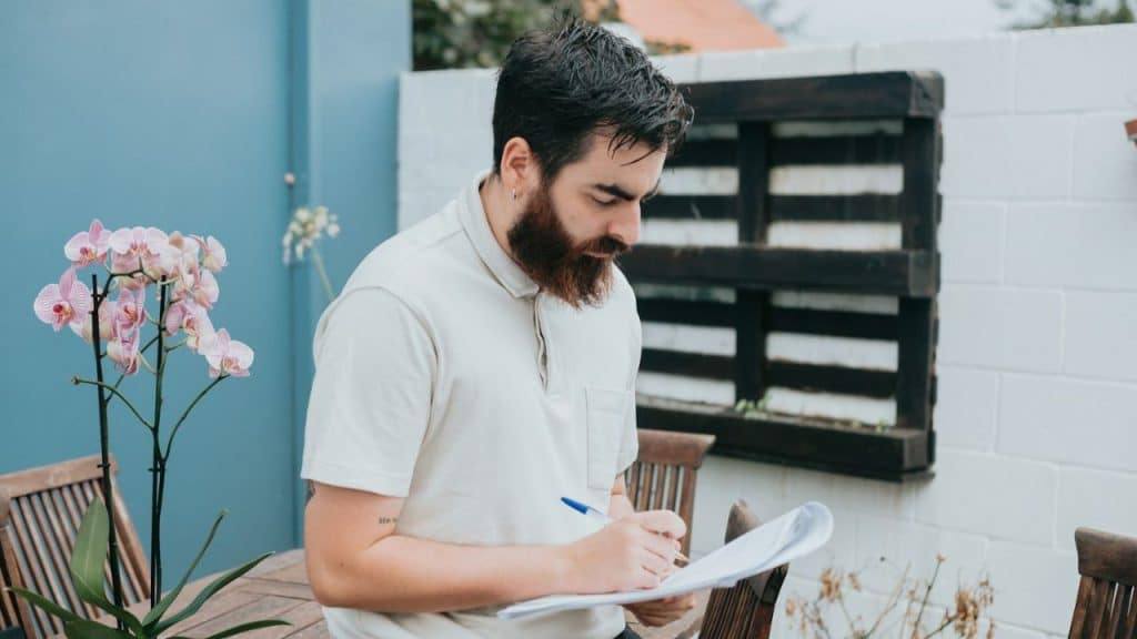 A bearded man in a light-colored polo shirt writes on a document outdoors.