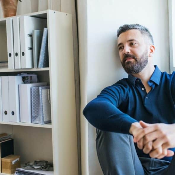 A pensive man with a beard leans against a wall next to a bookshelf.