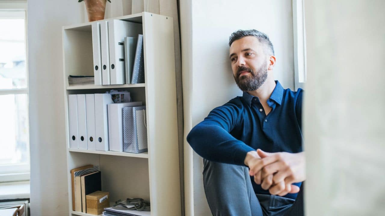A pensive man with a beard leans against a wall next to a bookshelf.