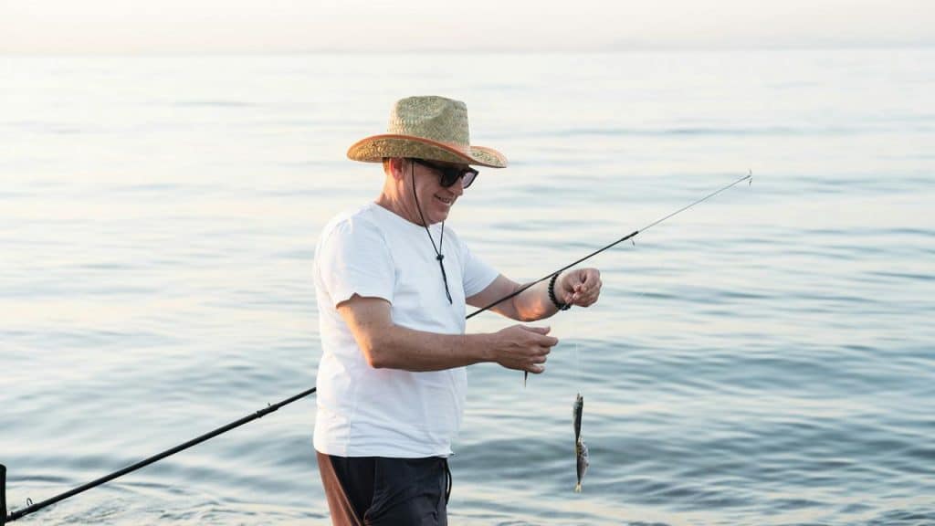 A man in a straw hat holding a small fish by the sea.