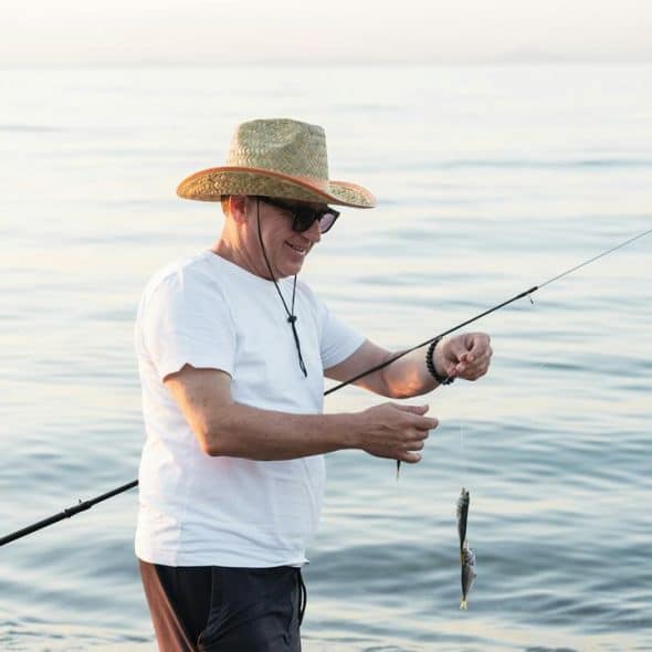 A man in a straw hat holding a small fish by the sea.