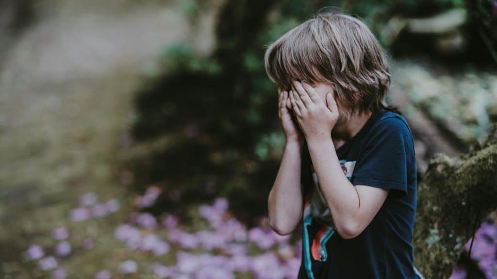 A boy in the woods covering his face with his hands.
