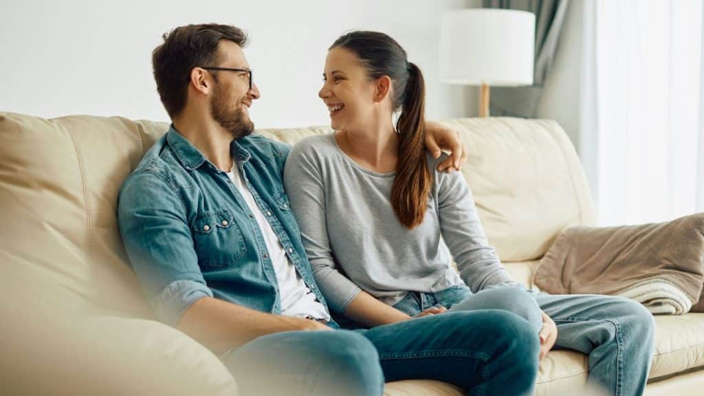 A cheerful couple sits closely on a beige couch, smiling at each other."